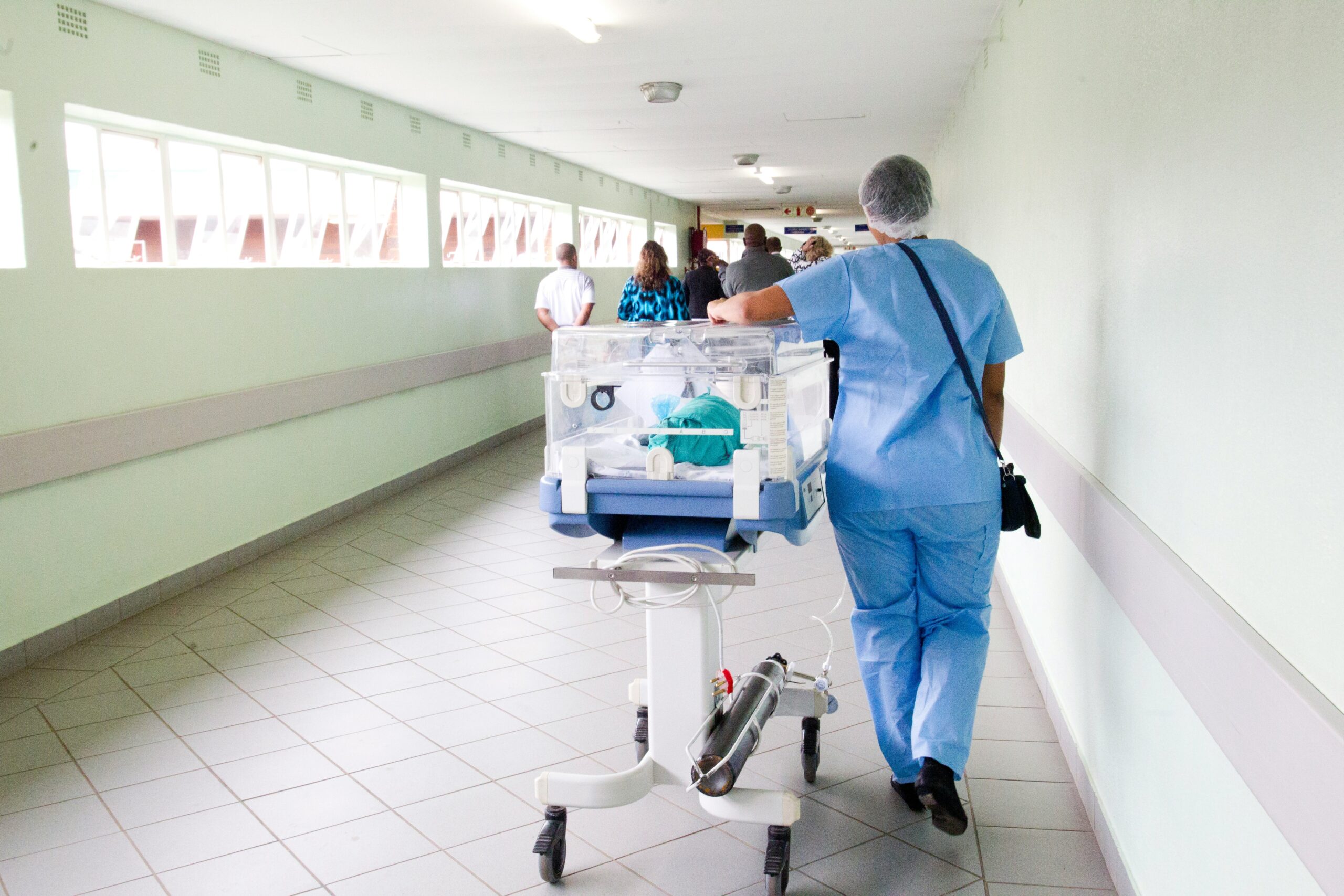 Healthcare professional in blue scrubs pushing a medical cart down a hospital corridor.