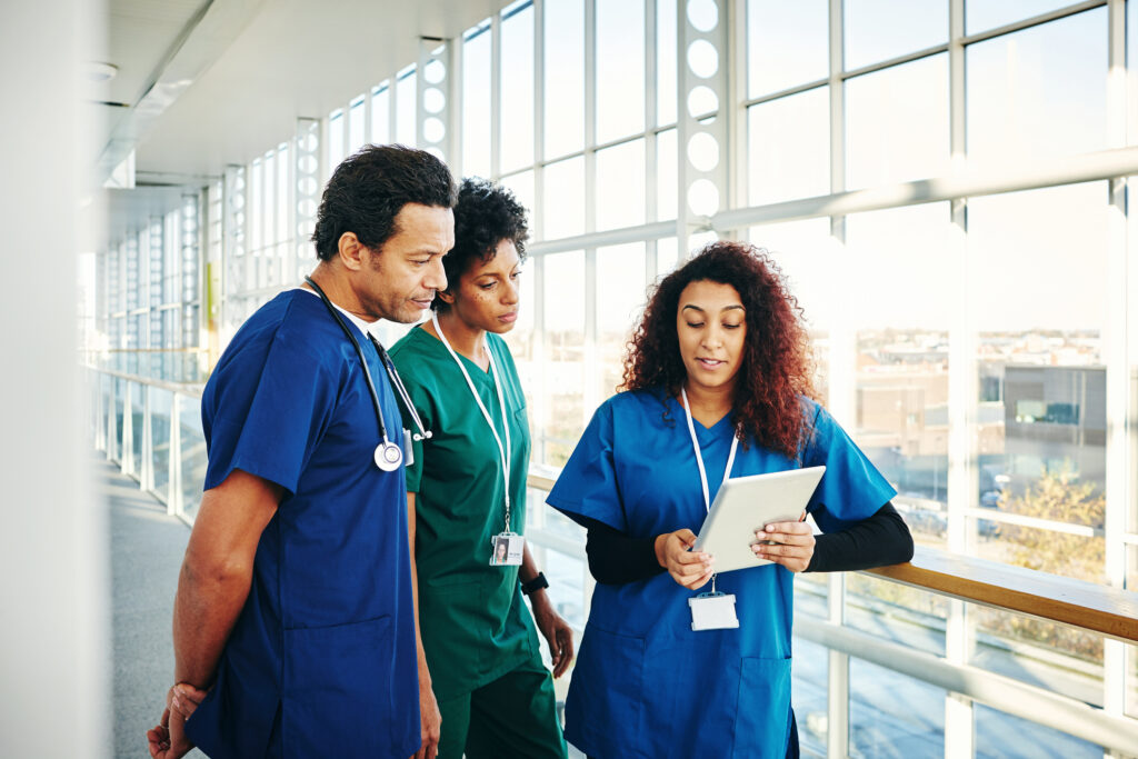 Three healthcare professionals in scrubs, consulting a digital tablet in a hospital walkway.