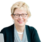 A smiling woman with short blonde hair, wearing glasses, a black sweater over a patterned blouse, and a silver necklace. She is sitting at a desk with a computer.